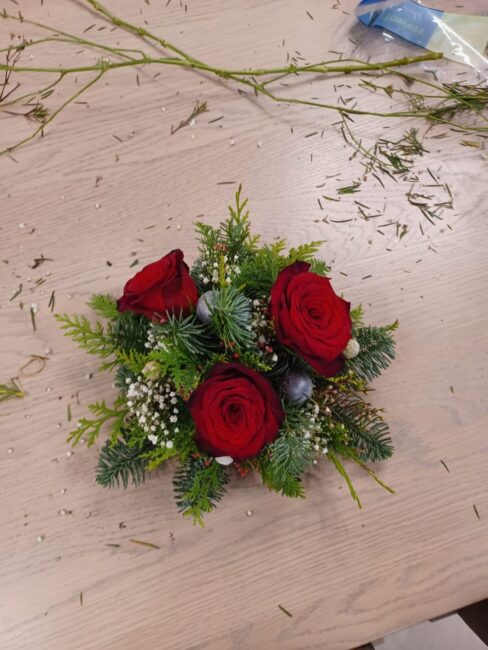Een bloemstuk met drie rode rozen, omringd door groene takken en witte bloemetjes, ligt op een houten tafel.