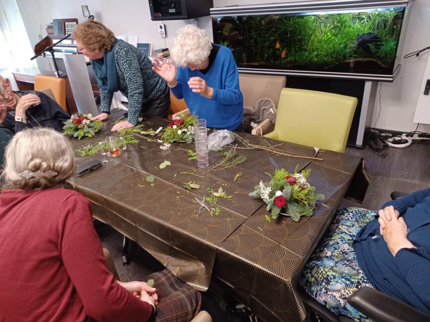 Een groep ouderen zit rond een tafel en maakt bloemstukken. Op de achtergrond is een groot aquarium zichtbaar.