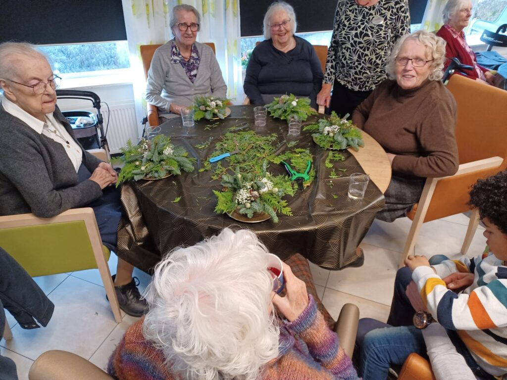 Een groep ouderen zit rond een tafel met groene planten en decoraties, glimlachend en in gesprek in een gezellige ruimte.