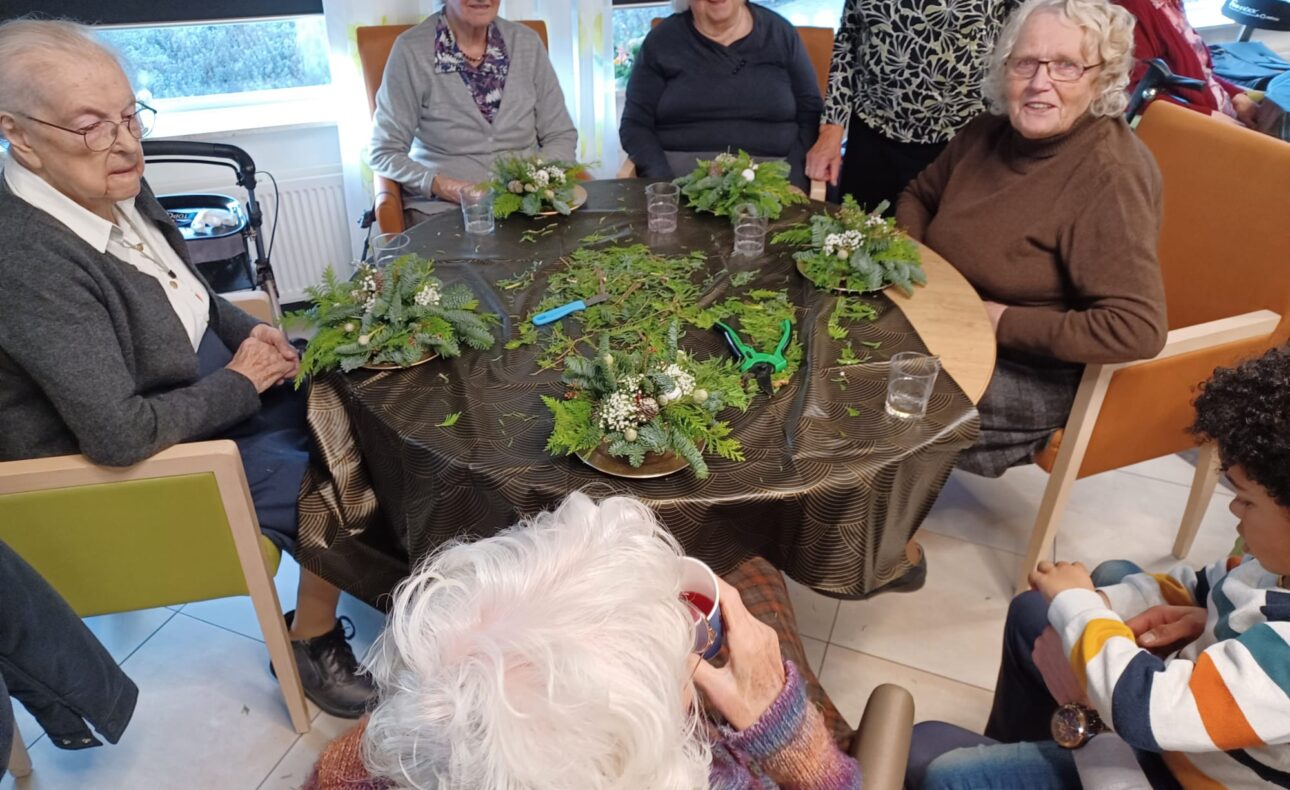 Een groep ouderen zit rond een tafel met groene planten en decoraties, glimlachend en in gesprek in een gezellige ruimte.
