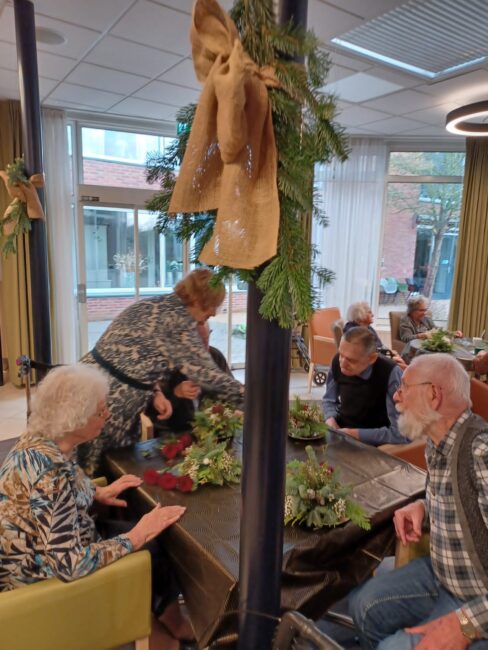 Een groep ouderen zit aan een tafel en maakt kerststukjes met groen en decoraties in een gemeenschappelijke ruimte.