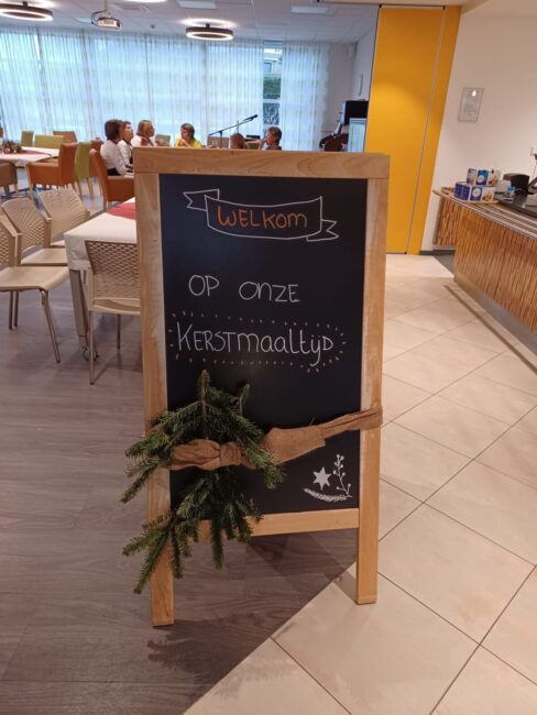Krijtbord in een eetzaal met de tekst "Welkom op onze Kerstmaaltijd", versierd met dennentakken en een strik.