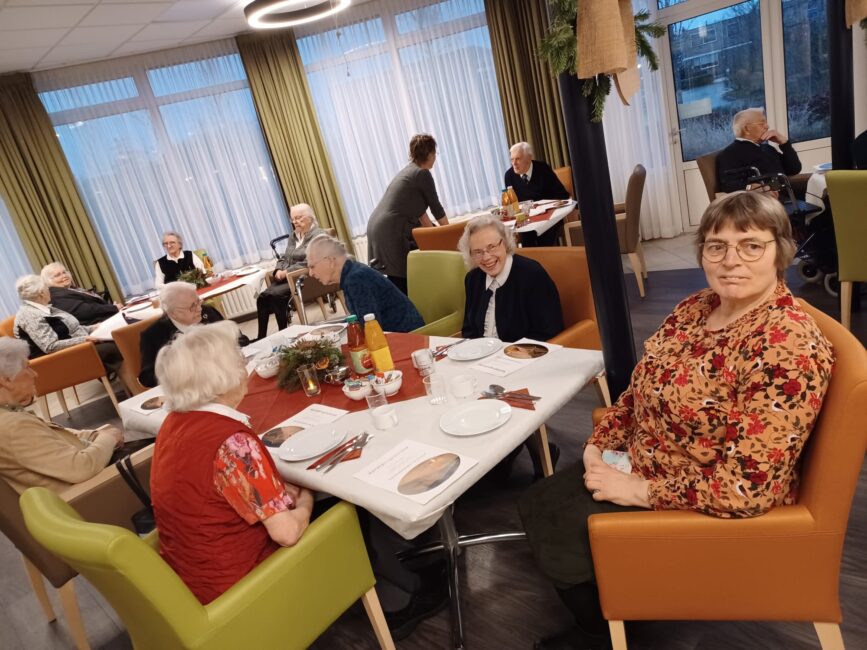 Groep ouderen zit aan een feestelijk gedekte tafel in een gezellige ruimte met grote ramen. Een persoon staat en bedient hen.