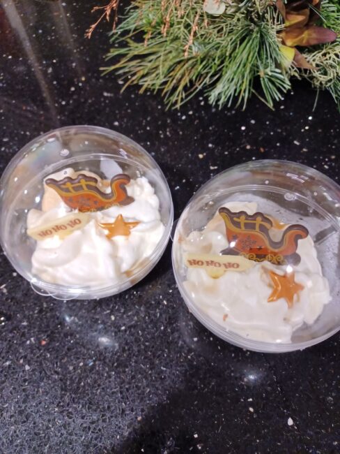 Twee plastic bakjes met slagroom en een kerstdecoratie op een zwart oppervlak, omringd door groene takken en kerstversiering.