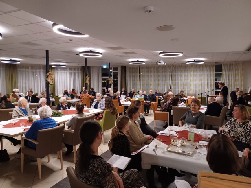 Groep mensen zit aan tafels in een verlichte zaal, gezellig pratend en etend. De ruimte is sfeervol ingericht met kerstdecoraties.