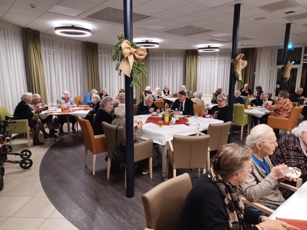 Een groep ouderen zit aan gedekte tafels in een sfeervol ingerichte zaal, versierd met kerstdecoraties en warme verlichting.