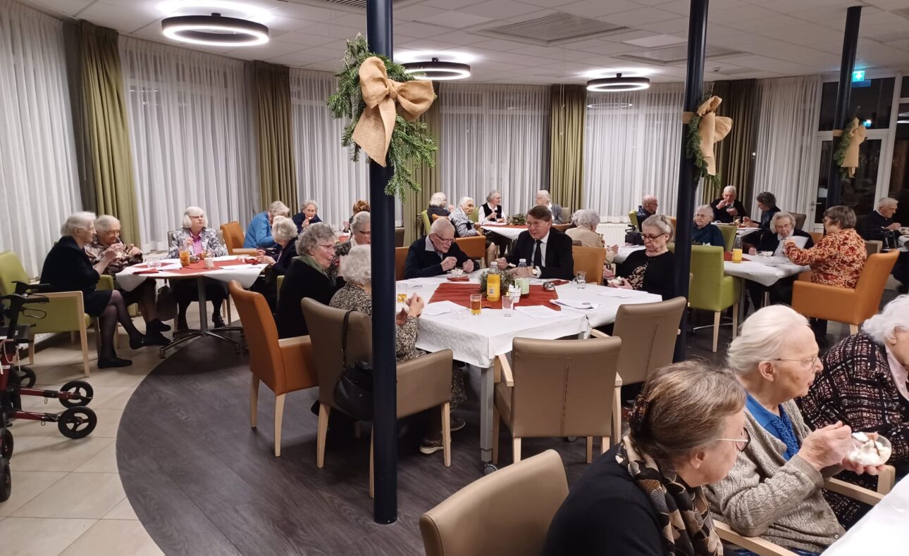 Een groep ouderen zit aan gedekte tafels in een sfeervol ingerichte zaal, versierd met kerstdecoraties en warme verlichting.