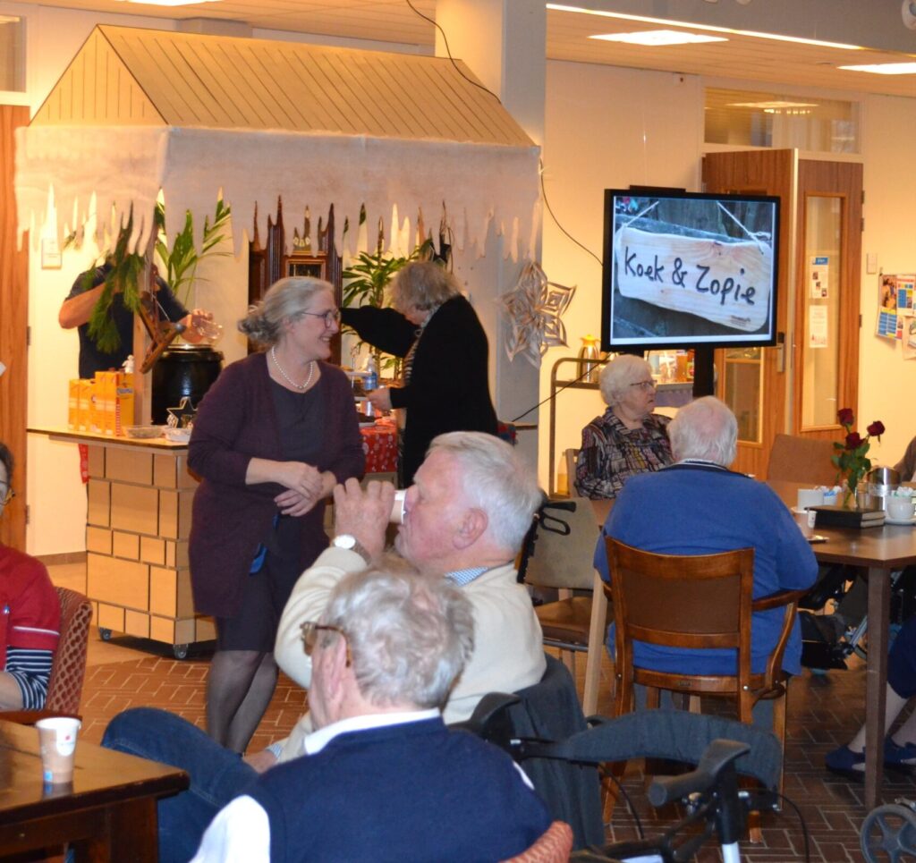 Mensen genieten van een gezellige bijeenkomst in een zaal met een koek-en-zopie kraam op de achtergrond.