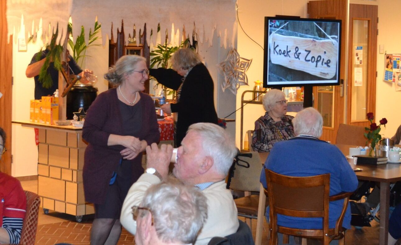Mensen genieten van een gezellige bijeenkomst in een zaal met een koek-en-zopie kraam op de achtergrond.