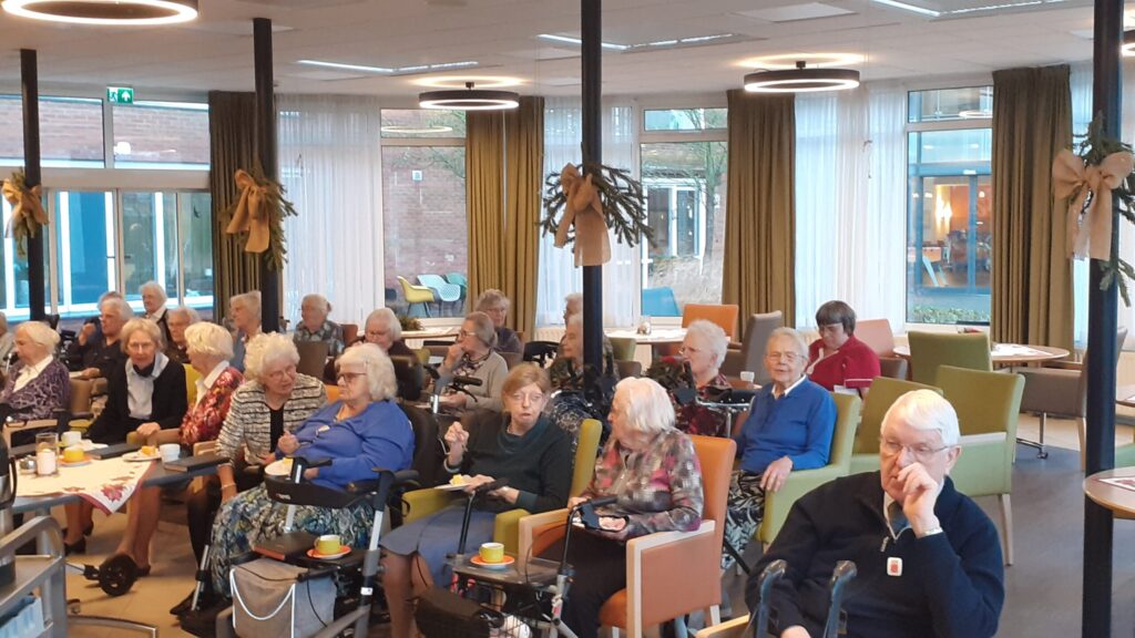 Groep ouderen zit in een gemeenschappelijke ruimte met tafels en stoelen, omringd door grote ramen en planten.