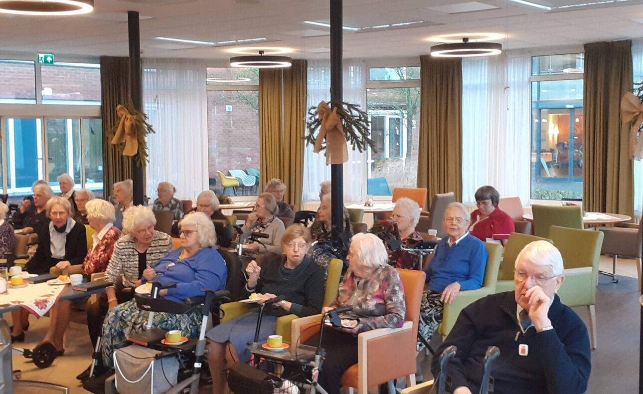 Groep ouderen zit in een gemeenschappelijke ruimte met tafels en stoelen, omringd door grote ramen en planten.