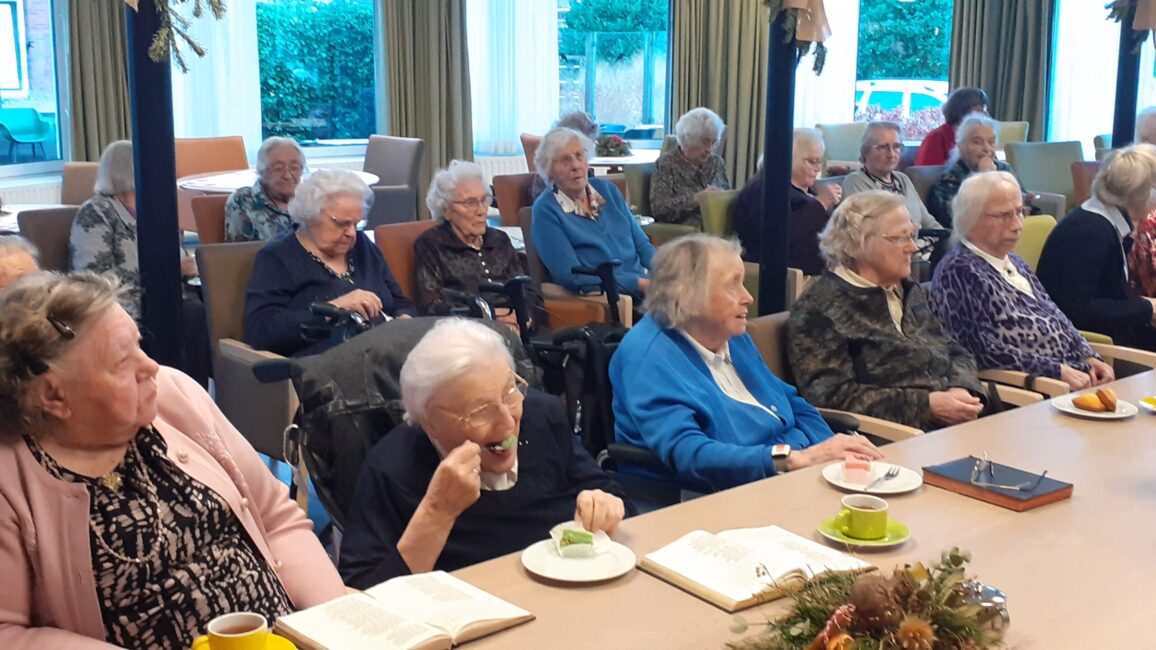Groep ouderen zit in een gemeenschappelijke ruimte, genietend van een activiteit. Tafels zijn gedekt met koffie en gebak.