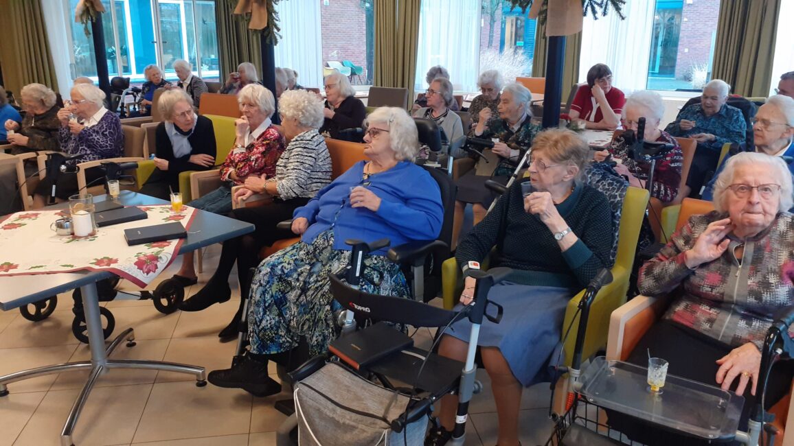 Groep oudere mensen zit in een gemeenschappelijke ruimte, gezellig pratend en genietend van drankjes in een verzorgingstehuis.