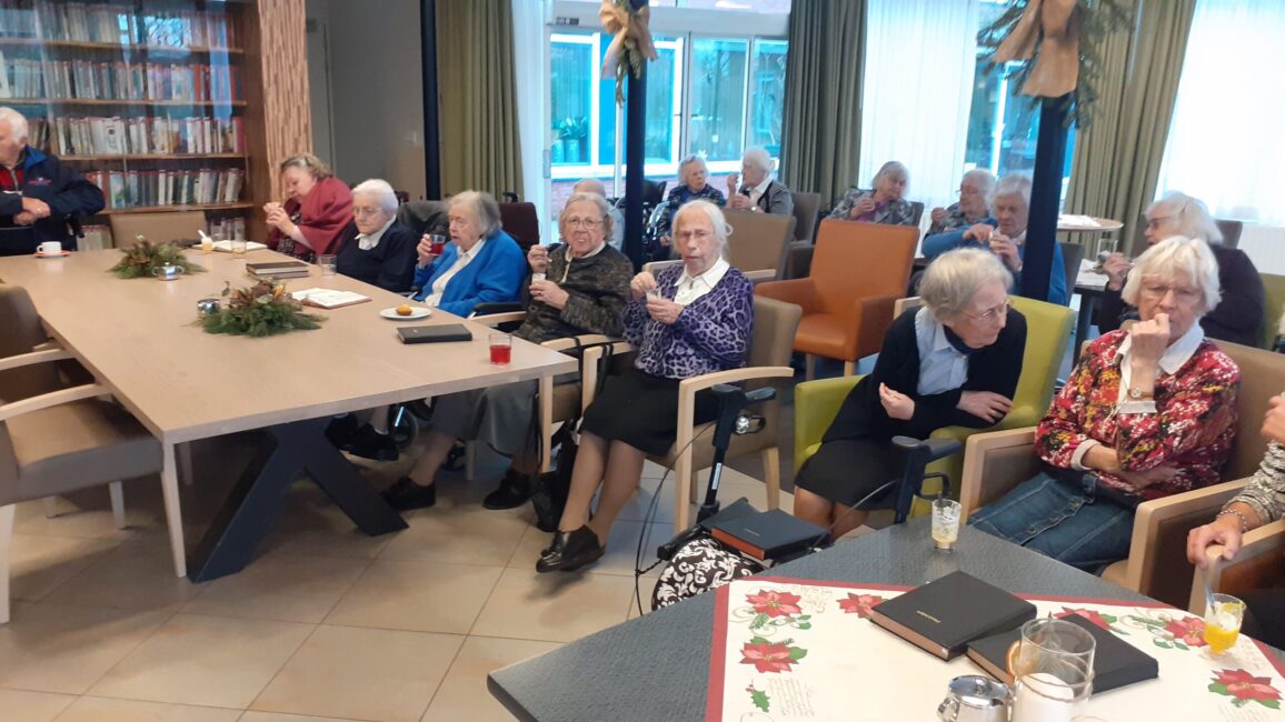 Groep ouderen zit in een gemeenschappelijke ruimte, omringd door boekenplanken en kerstdecoraties, in gesprek met elkaar.