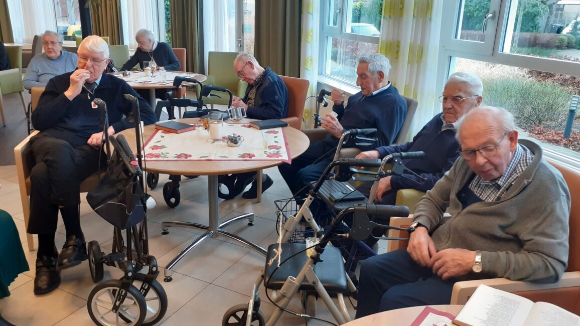 Een groep ouderen zit in een gemeenschappelijke ruimte van een verzorgingstehuis. Sommigen lezen, anderen rusten.