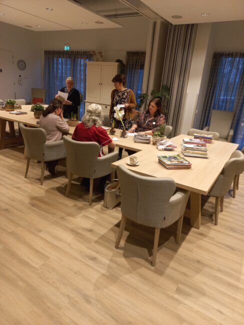 Een groep mensen zit aan een tafel in een gezellige ruimte met houten vloer, omringd door boeken en planten.