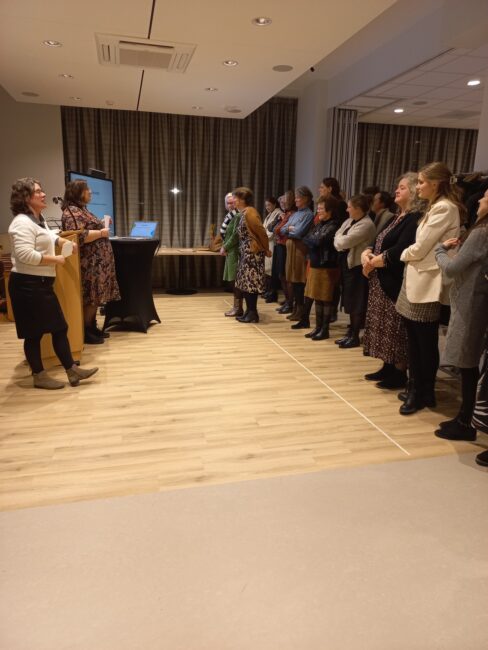 Een groep mensen staat in een zaal, aandachtig luisterend naar een presentatie. Twee personen staan vooraan bij een scherm.