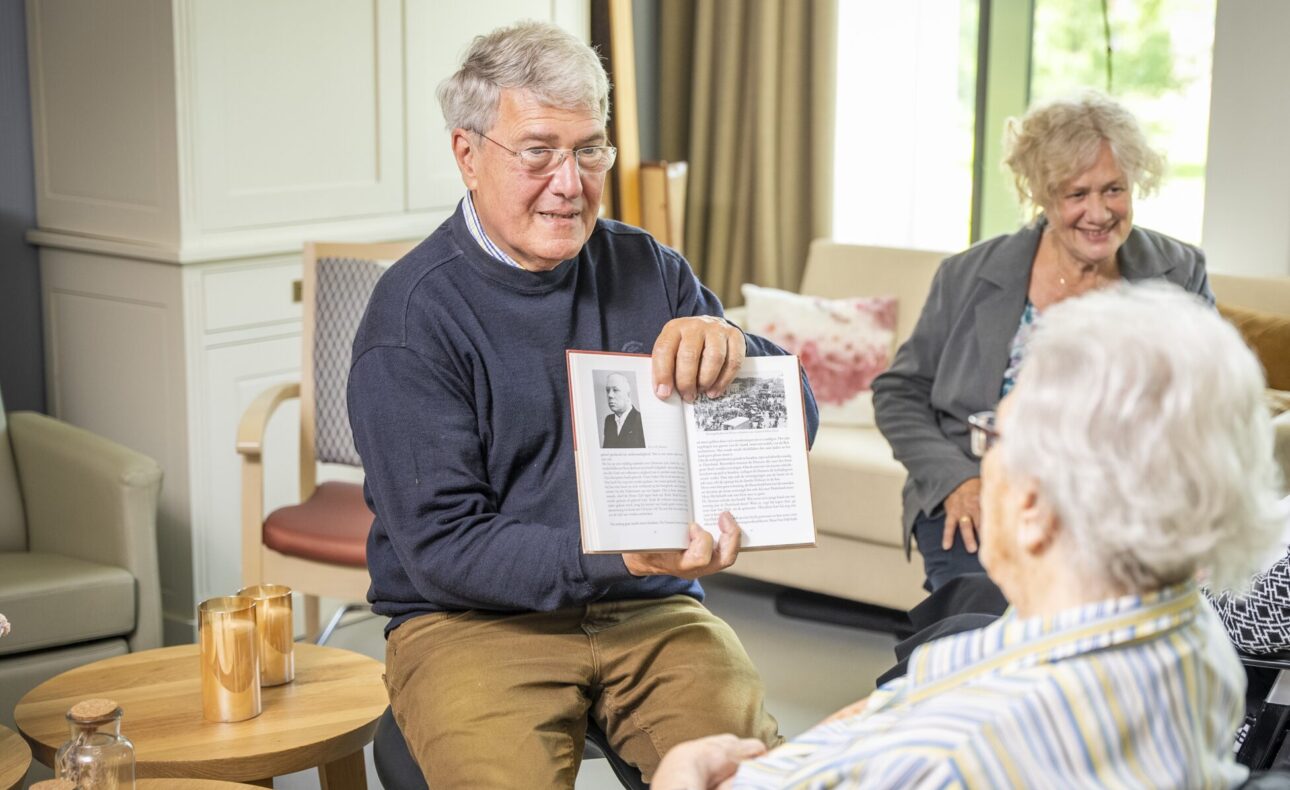 Een oudere man leest voor uit een boek aan een groep senioren in een gezellige woonkamer. Iedereen luistert aandachtig.