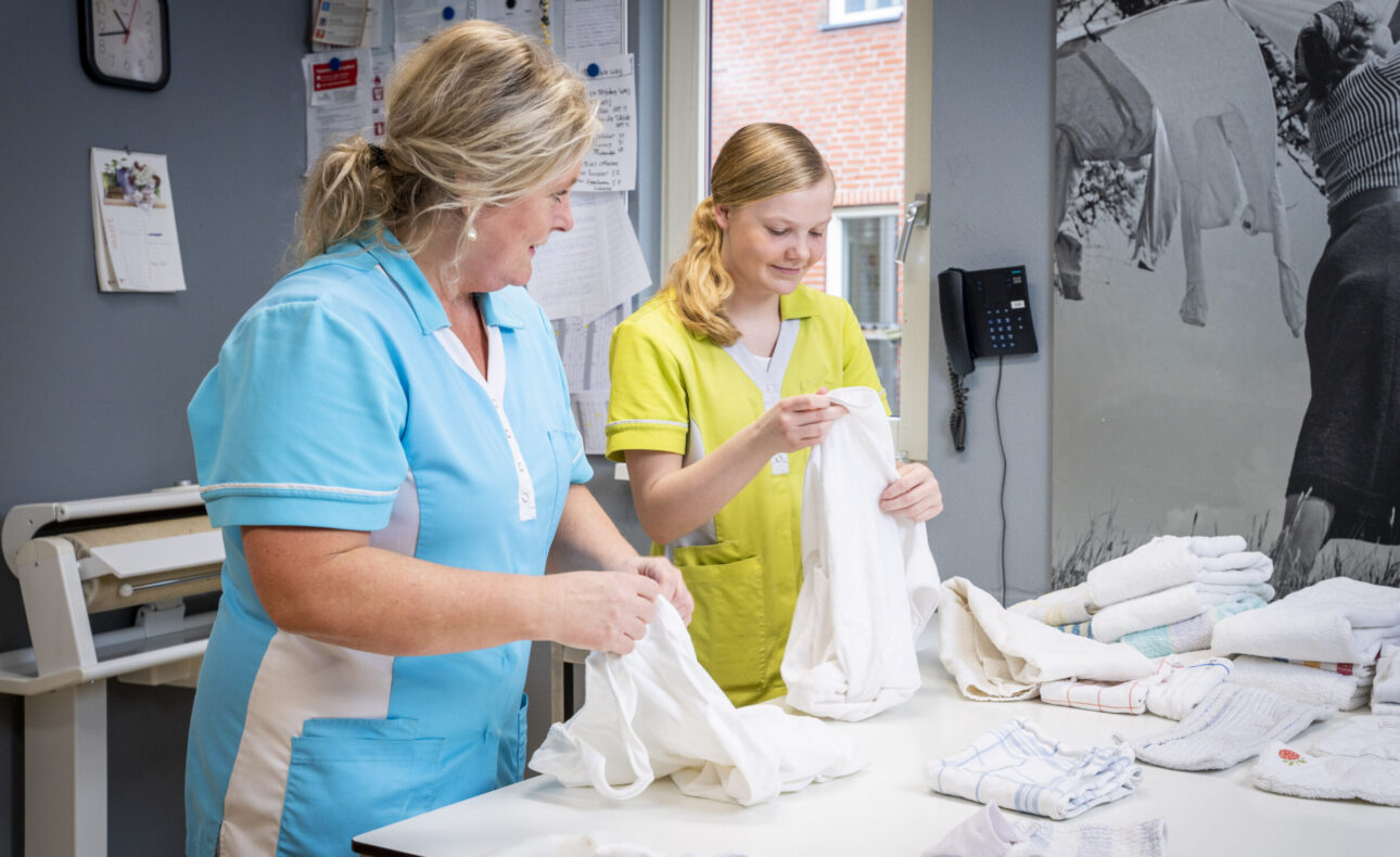 Twee zorgmedewerkers in uniform vouwen wasgoed in een lichte kamer met een raam en muurdecoratie op de achtergrond.