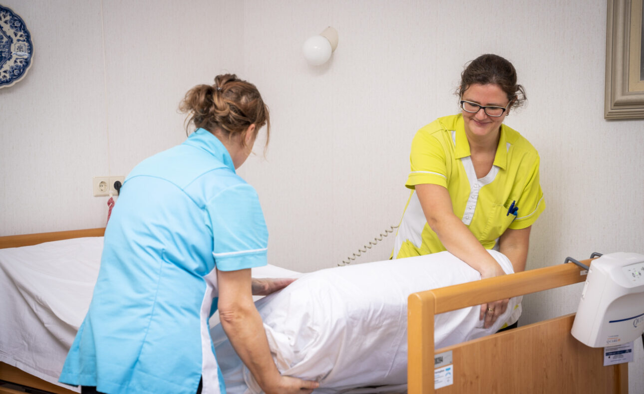 Twee zorgmedewerkers maken samen een bed op in een zorginstelling, gekleed in blauwe en gele uniformen.