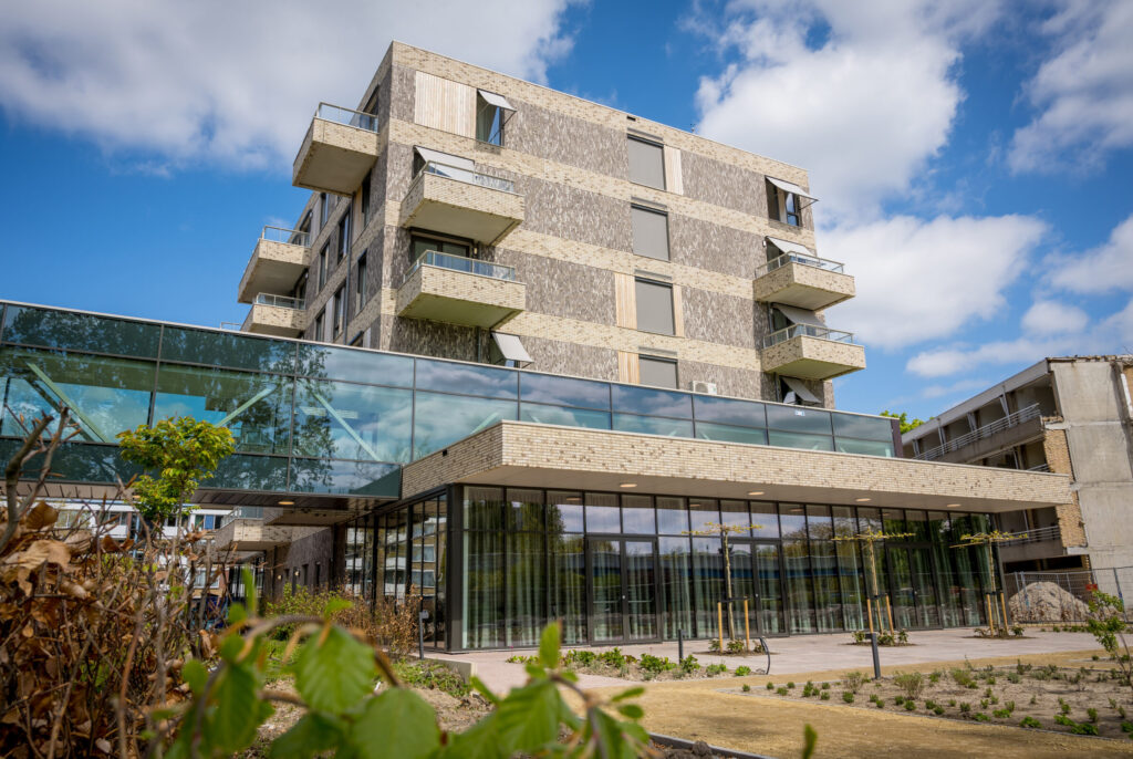 Modern gebouw met glazen gevel en meerdere verdiepingen, omgeven door groen en een heldere blauwe lucht met enkele wolken.