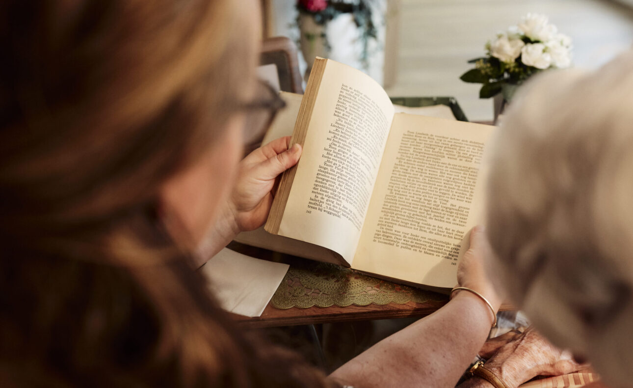 Twee mensen lezen samen een oud boek aan een tafel, met een vaas bloemen op de achtergrond. Hun gezichten zijn niet zichtbaar.