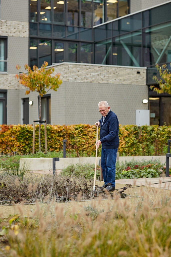 Een oudere man werkt met een hark in een tuin voor een modern gebouw, omringd door herfstkleuren en struiken.