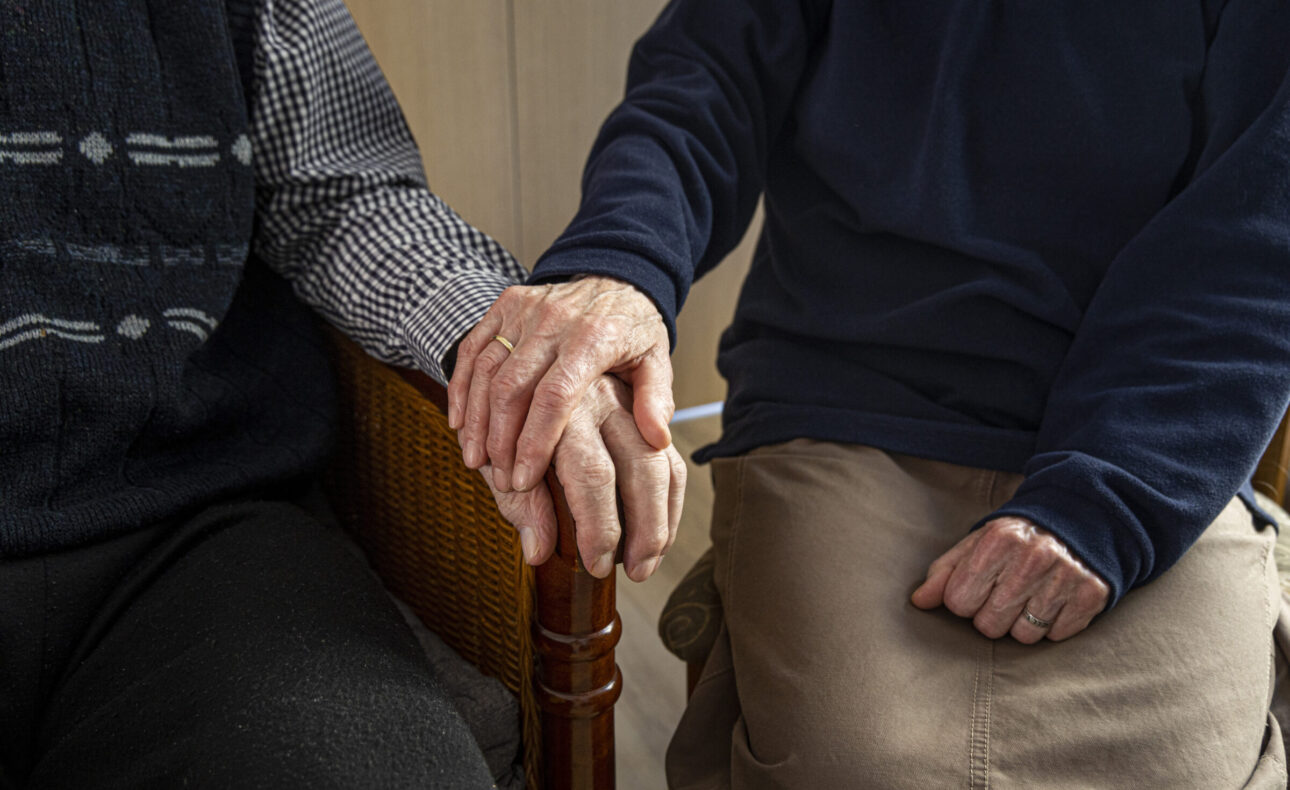 Twee oudere mensen zitten naast elkaar en houden elkaars handen vast, zittend op stoelen in een warme, huiselijke omgeving.