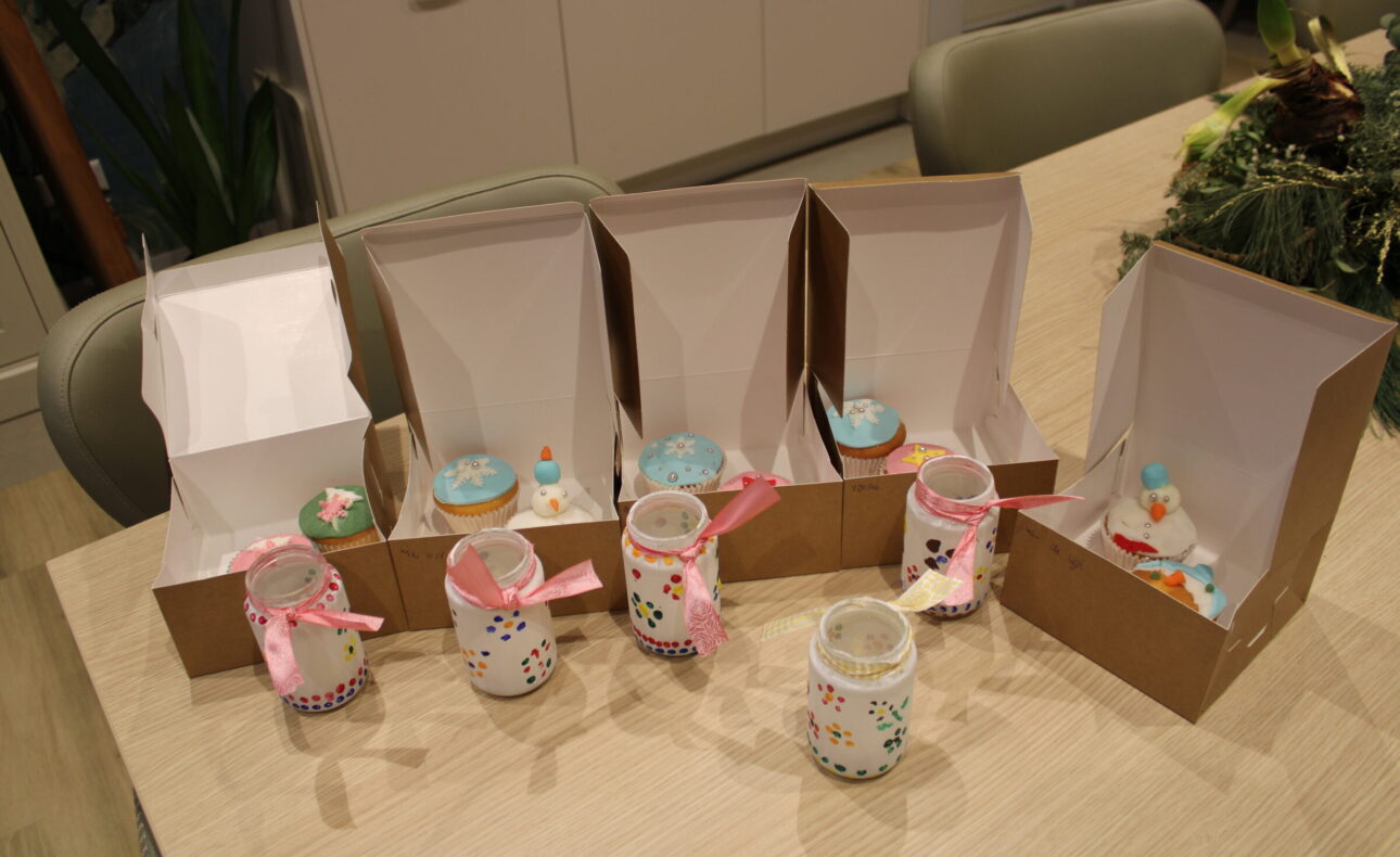 Een tafel met versierde cupcakes in open dozen en glazen potjes met roze linten, klaar voor een feestelijke gelegenheid.