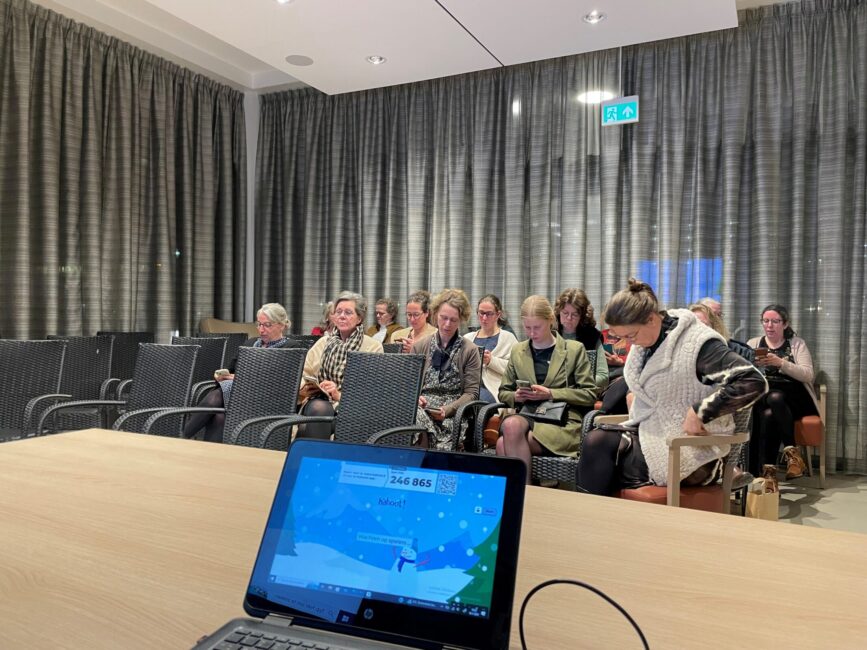 Een groep mensen zit in een zaal met stoelen, kijkend naar een scherm. Op de voorgrond is een laptop zichtbaar op een tafel.