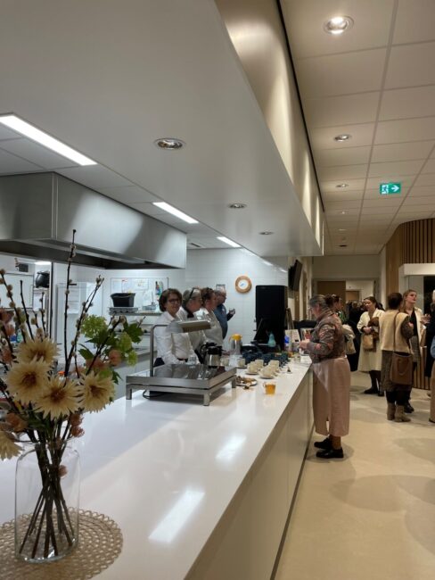 Een moderne keuken met mensen die praten en eten. Op de voorgrond staat een vaas met bloemen op een witte balie.