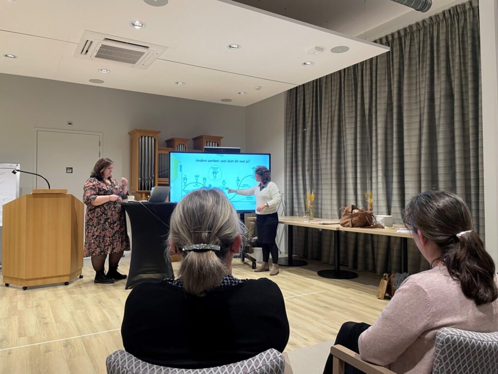 Een presentatie in een vergaderruimte met een groot scherm. Twee mensen staan vooraan, terwijl anderen toekijken vanaf stoelen.
