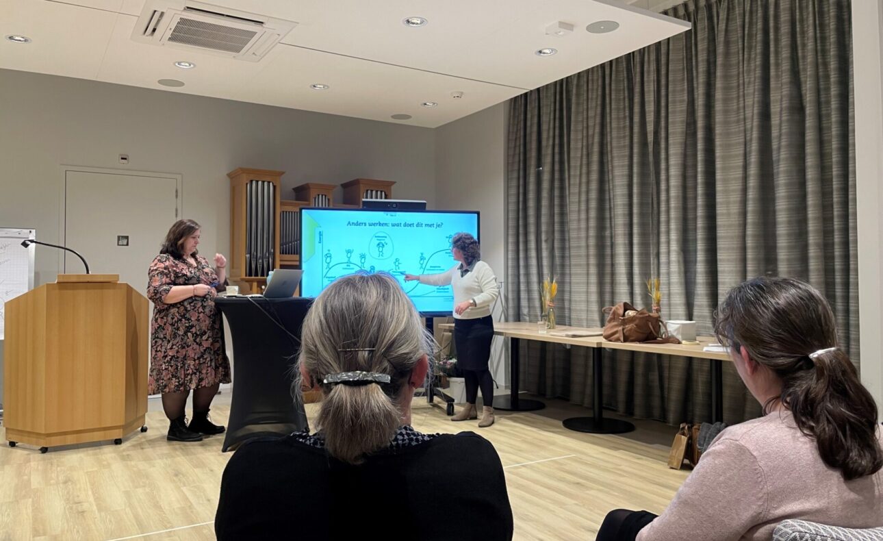 Een presentatie in een vergaderruimte met een groot scherm. Twee mensen staan vooraan, terwijl anderen toekijken vanaf stoelen.