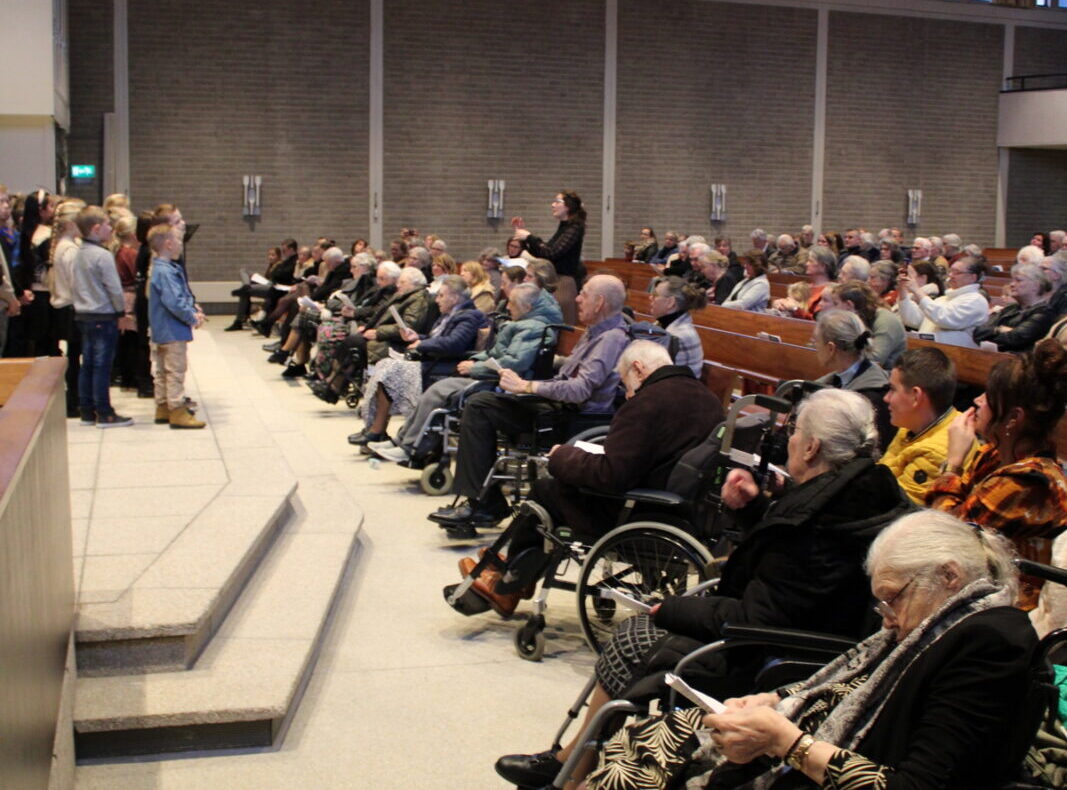 Een groep mensen, waaronder ouderen in rolstoelen, zit in een zaal met hoge ramen en houten plafond, kijkend naar een podium.
