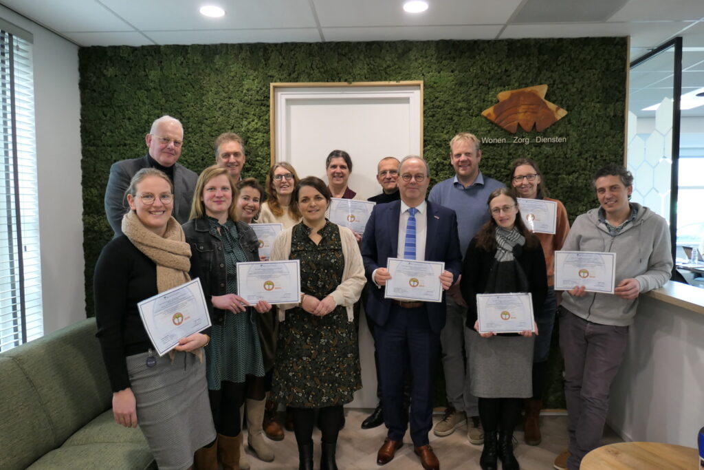 Groep mensen poseert met certificaten in een kantoor met groene muur. Ze lachen en staan dicht bij elkaar voor een foto.