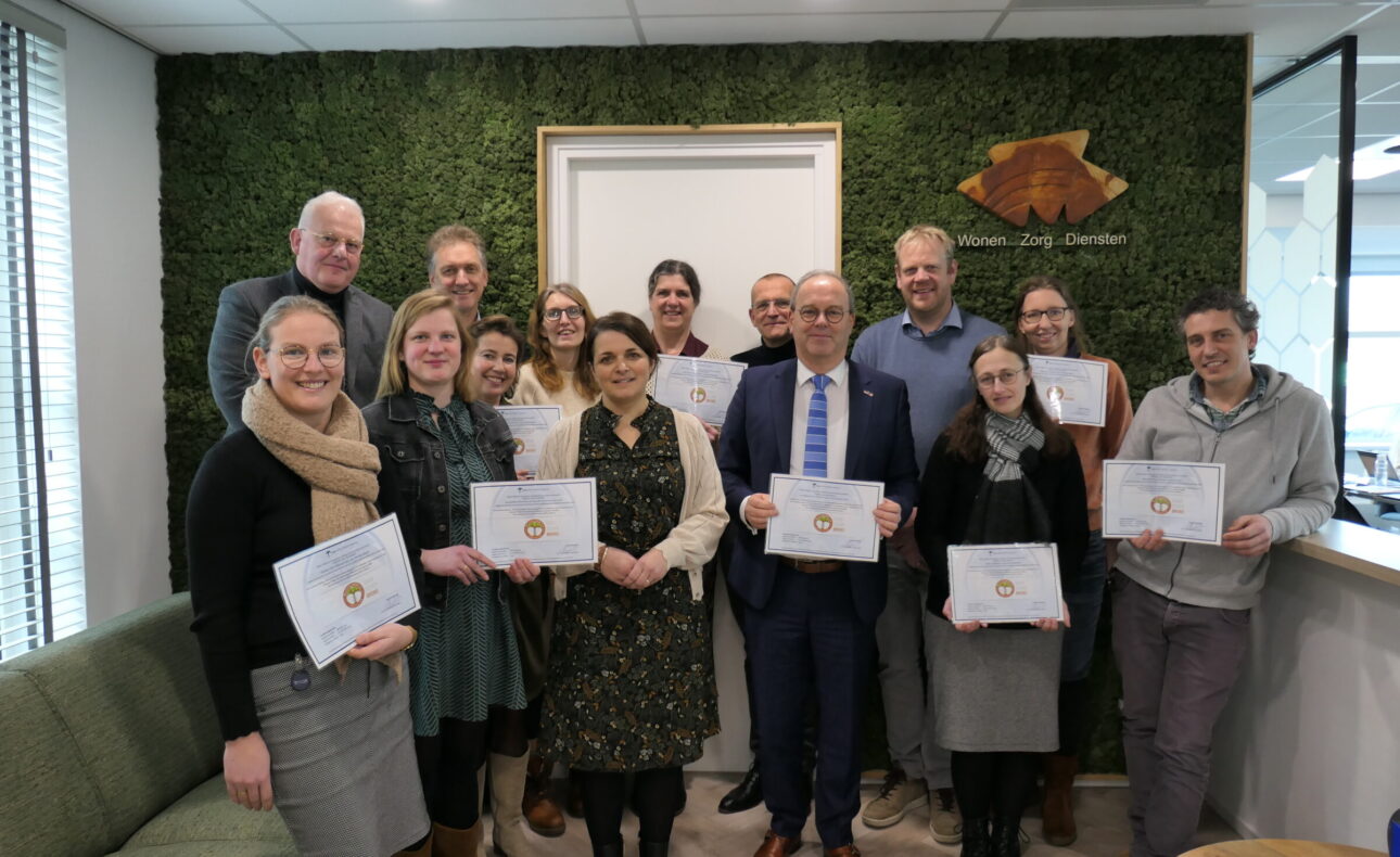 Groep mensen poseert met certificaten in een kantoor met groene muur. Ze lachen en staan dicht bij elkaar voor een foto.