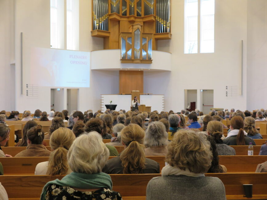 Een volle zaal met mensen die luisteren naar een spreker tijdens een congres over palliatieve zorg in een kerkachtige ruimte.