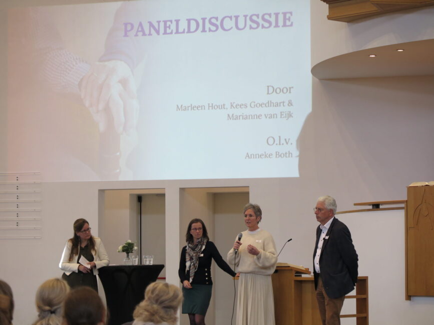 Een paneldiscussie over palliatieve zorg met vier sprekers op een podium, met een projectiescherm op de achtergrond.