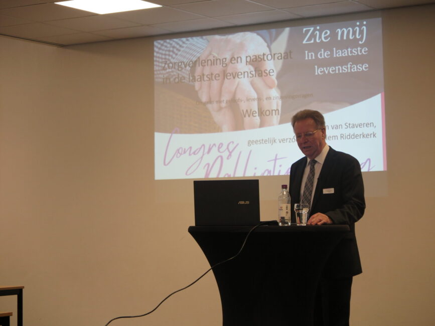 Man geeft presentatie over palliatieve zorg op een congres, met een projectiescherm en een laptop op een tafel.