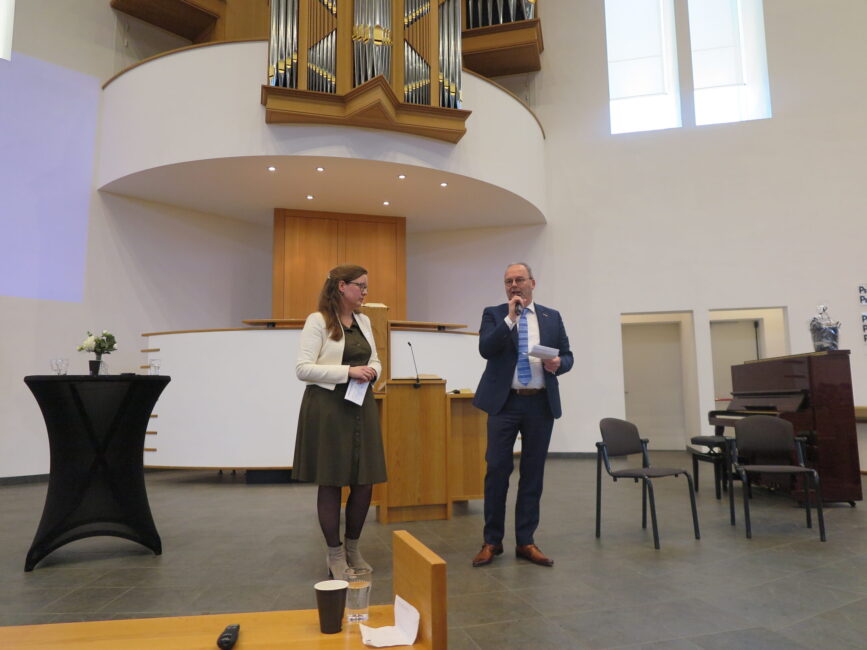 Twee mensen staan op een podium in een moderne kerkzaal, met een orgel op de achtergrond, tijdens een presentatie of congres.
