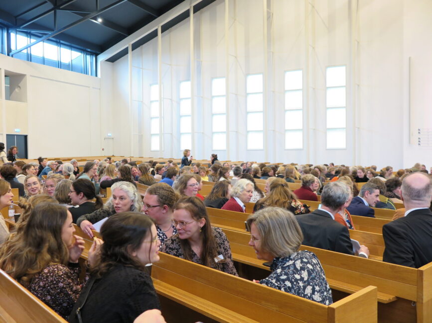 Een volle zaal met mensen die op houten banken zitten tijdens een congres over palliatieve zorg, met grote ramen op de achtergrond.