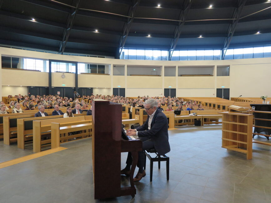 Een man speelt piano in een grote zaal met publiek, tijdens een congres over palliatieve zorg. De zaal is modern ingericht.