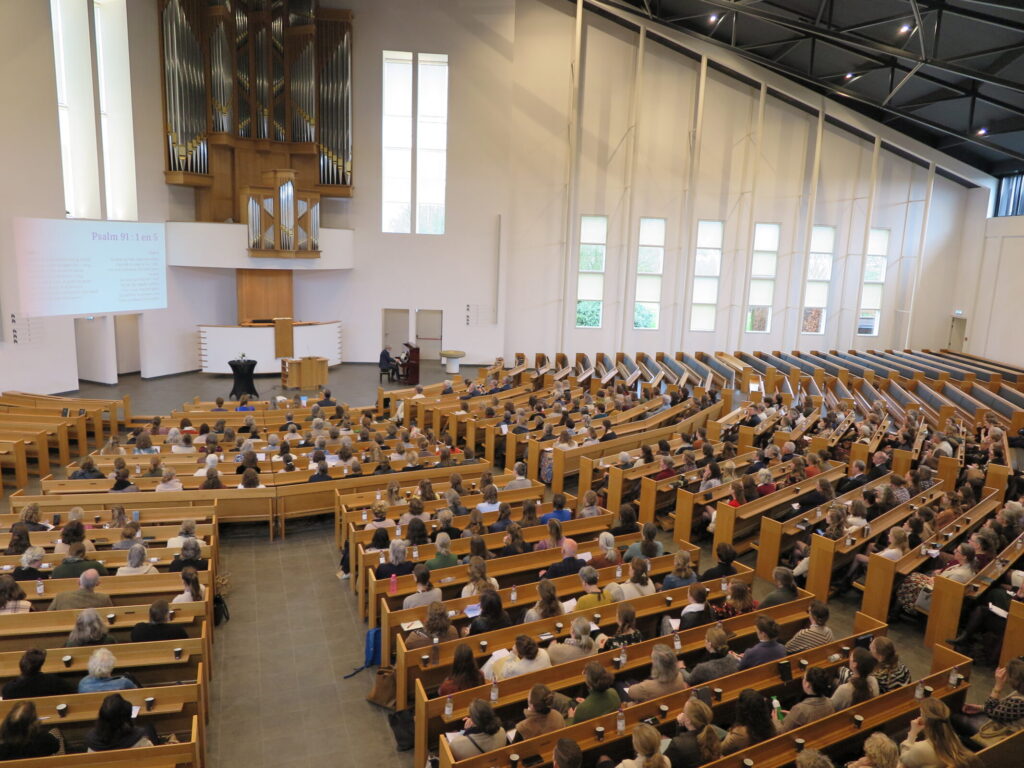 Een volle conferentiezaal met houten banken en een groot orgel aan de muur, waar een presentatie over palliatieve zorg plaatsvindt.