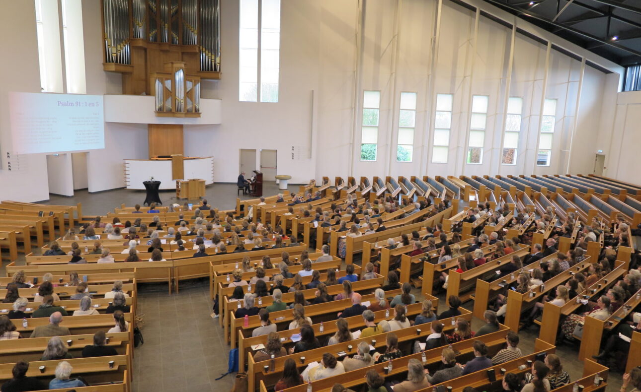 Een volle conferentiezaal met houten banken en een groot orgel aan de muur, waar een presentatie over palliatieve zorg plaatsvindt.