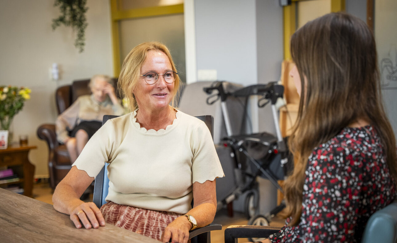 Twee vrouwen zitten aan een tafel in een gezellige kamer. Op de achtergrond is een oudere persoon in een stoel te zien.