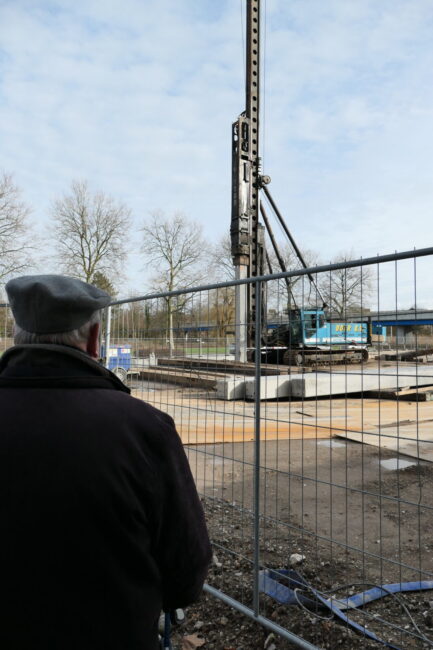 Een oudere man kijkt naar een bouwplaats met een grote heistelling achter een hek op een bewolkte dag.