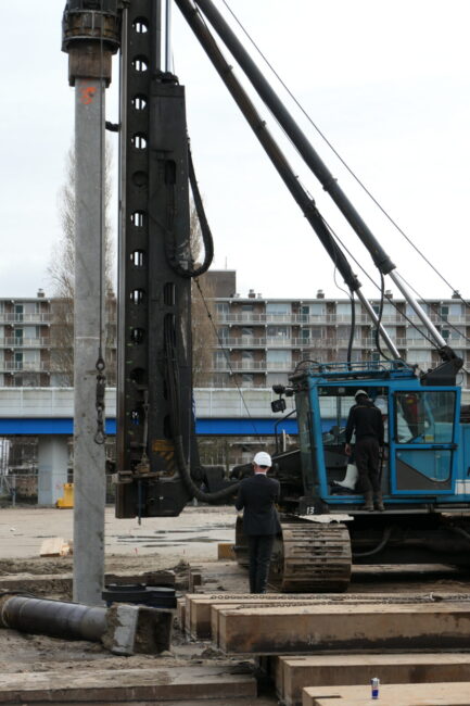 Een bouwplaats met een grote heistelling en arbeiders in veiligheidshelmen. Op de achtergrond zijn flats zichtbaar.