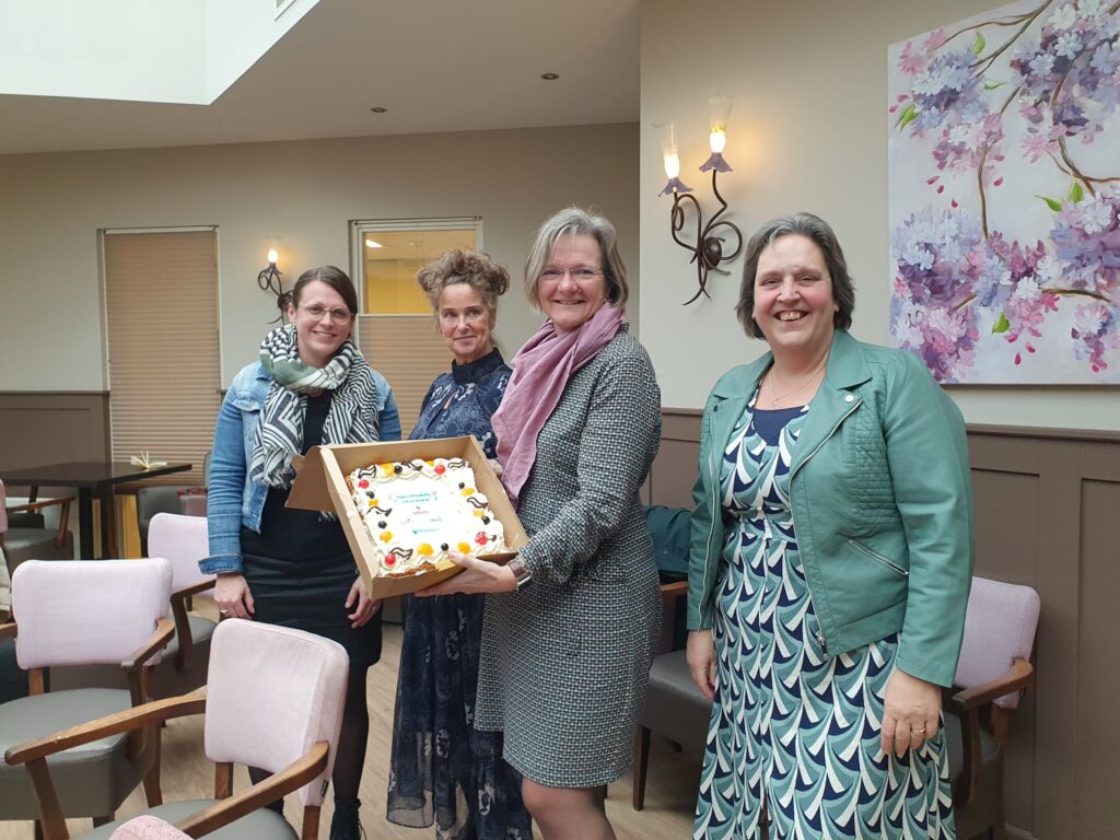 Vier vrouwen poseren glimlachend met een taart in een gezellige kamer met schilderijen en zachte stoelen op de achtergrond.