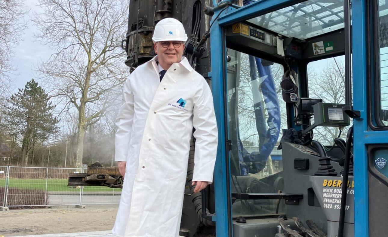Man in witte jas en helm staat op een bouwvoertuig. Hij draagt witte laarzen. Op de achtergrond zijn bomen zichtbaar.