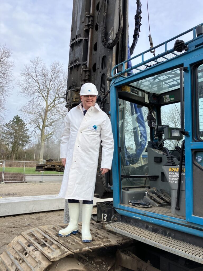Man in witte jas en helm staat op een bouwvoertuig. Hij draagt witte laarzen. Op de achtergrond zijn bomen zichtbaar.